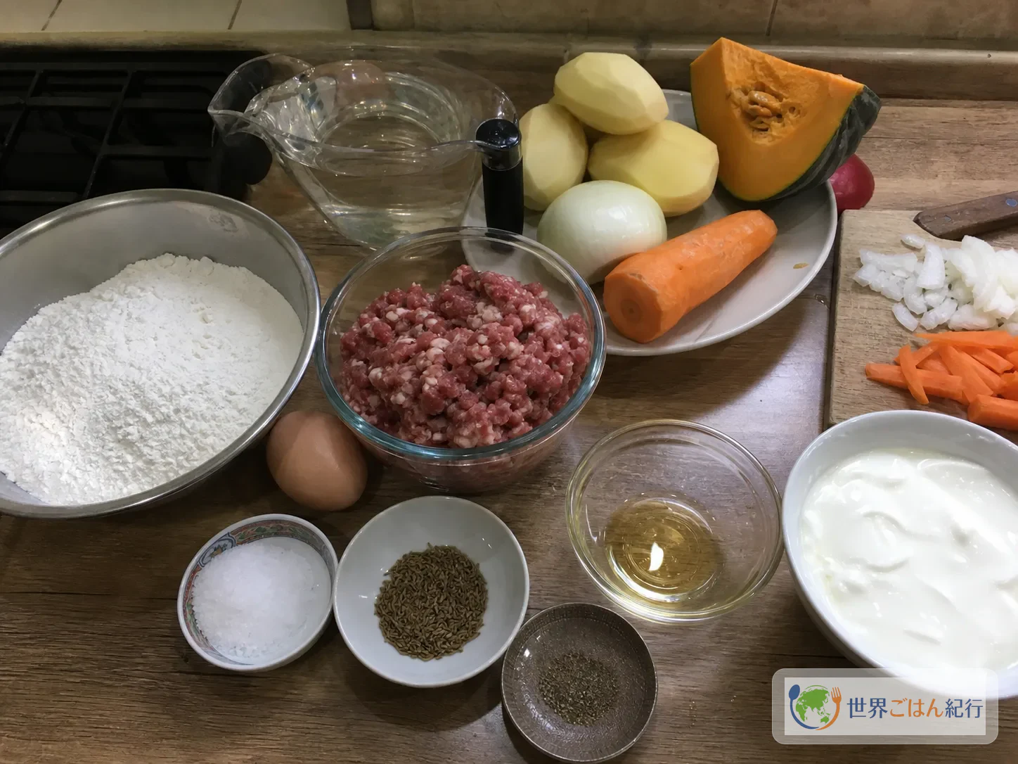 オロモの材料。粉、卵、牛ひき肉、じゃがいも、玉ねぎ、にんじん、かぼちゃ、クミン、ヨーグルトを並べた様子